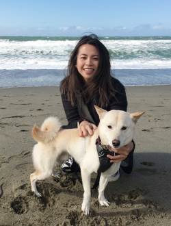 A woman with long dark hair crouching next to a light colored dog on the beach with the ocean in the background