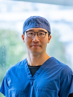 Man with glasses wearing blue scrubs standing in front of a large window