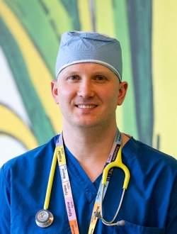 Man smiling wearing blue scrubs with a stethoscope around his neck and a green background