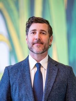 Man with short dark wavy hair and a black and white bear wearing a blue suit and tie with a green background