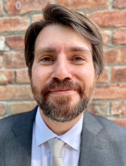 Man with a brown beard and hair wearing a suite and tie standing in front of a brick wall