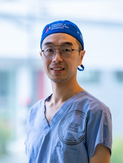 Man with glasses standing in front of a window wearing blue scrubs and a blue cap