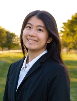 Smiling woman with long black hair wearing a white shirt and dark jacket with green trees and lawn in the background