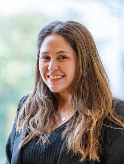 Smiling woman with long brown hair wearing a dark top and jacket standing in front of a window