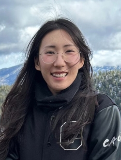 Smiling woman with glasses and long black hair, wearing a letter jacket with Emerald Bay with storm clouds in the background