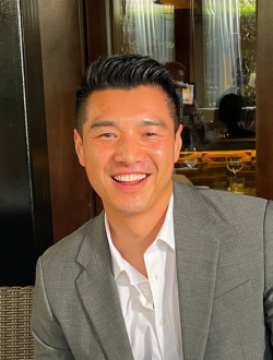 Smiling man with short black hair wearing a grey suit jacket over a shite shirt