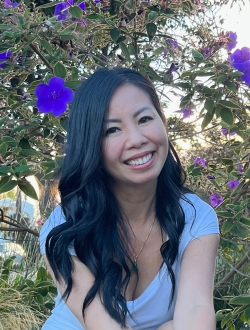 Woman with long black hair smiling, with a flowering plant in the background