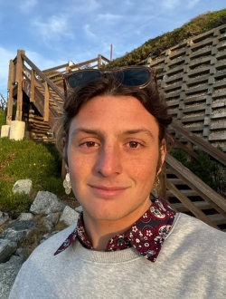 Man with long brown hair pulled back, wearing a grey sweater with a cloudy sky in the background