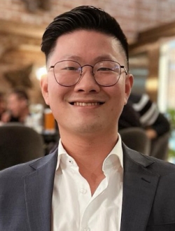 Man smiling with glasses and short black hair wearing a grey blazer over a white shirt open at the neck