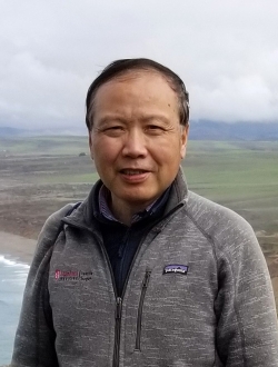 Man smiling with short black hair wearing a gray sweater with a cloudy coastal scene in the background