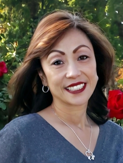 Woman with long dark brown hair smiling and wearing a gray sweater with roses and greenery in the background.