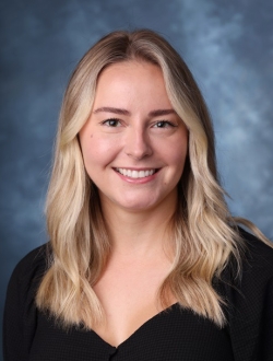 Woman with long blond hair smiling, wearing a dark top in front of a blue background.