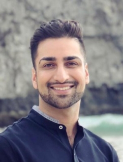 Smiling man with short dark hair and beard with a cliff and water in the background.