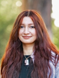 Woman with long brown hair with greenery in the background.