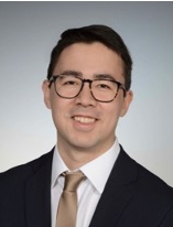 Smiling man with short black hair and glasses wearing a suit and tie.