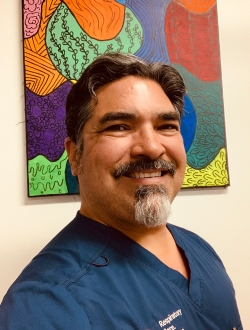 Man with dark hair and a salt and pepper goatee wearing blue scrubs.