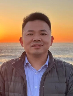 Smiling man with short black hair wearing a gray jacket over a blue button down shirt with the sun setting over the ocean in the background.