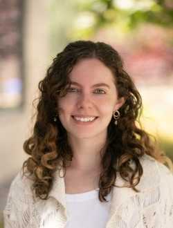 Woman with long curly brown hair wearing a cream sweater over a white shirt with leafy greenery in the background.