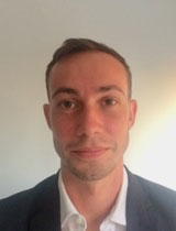 Man with short brown hair wearing a gray suit jacket over a while collared shirt.