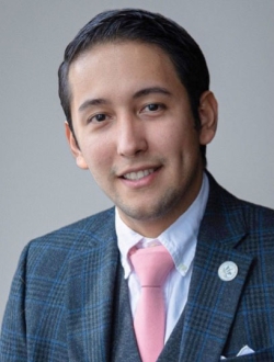 Smiling man with short black hair, wearing a gray and blue suit with a pink tie.
