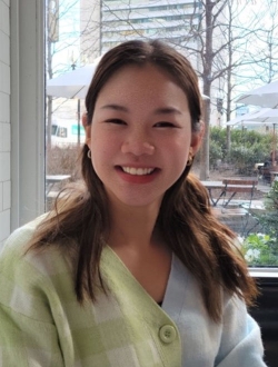 Woman with long brown hair wearing a light blue and green sweater and sitting in front of a window with a view of a cityscape.