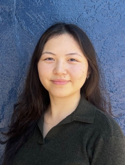 Woman with long black hair wearing a black shirt in front of a dark blue textured wall.