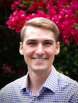 Smiling man with short blond hair wearing a collared shirt with red flowers in the background.
