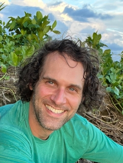 Smiling man in with wavy brown hair wearing a green shirt with greenery and clouds in the background.