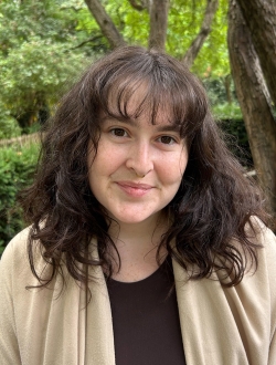 Woman with long wavy dark brown hair wearing a beige sweater with trees in the background.