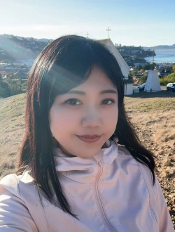 Smiling woman with long straight dark brown hair wearing a pink windbreaker with hills, houses, and water in the background.