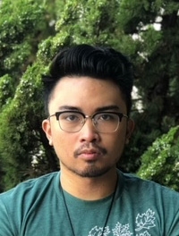 Man with short black hair wearing glasses and a green shirts in front of a natural green background.