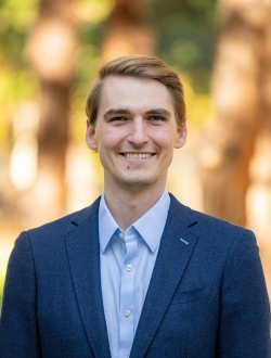 Smiling man with short blond hair wearing a collared shirt and blue suit jacket with trees in the background..