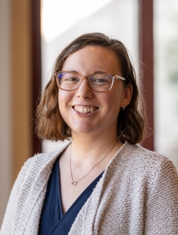 Smiling woman with glasses and shoulder length wavy brown hair, wearing a light sweater over a dark blue top.