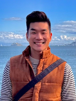 Man with short black hair, wearing an orange vest with a background of water, sky and clouds.