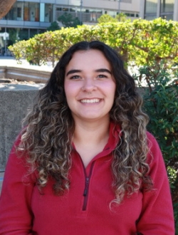 Woman with long curly brown hair wearing a red sweater with trees, building and bushes in the background.