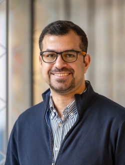 Smiling man with short dark hair and beard wearing glasses and a blue sweater over a striped collared shirt.