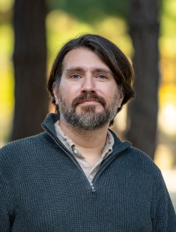 Man with medium long brown hair and a salt and pepper beard wearing a sweater with trees in the background.