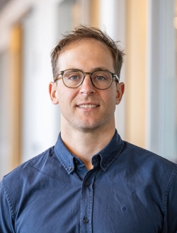 Smiling man with short brown hair wearing glasses and a blue button downed shirt.