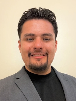 Man with short black hair and a goatee beard wearing a gray suit jacket over a black t-shirt.