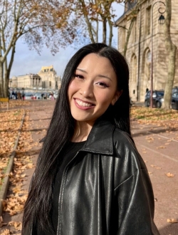 Smiling woman with long black hair wearing a black coat with a tree-lined avenue in the background.
