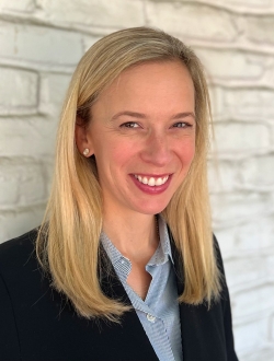 Smiling woman with long blonde hair wearing a blue shirt and navy blazer standing in front of a white brick wall.