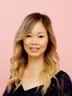 Woman with long light brown hair wearing a dark top in front of a pink background.