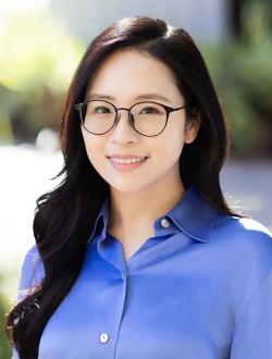 Smiling woman with long dark hair wearing glasses and a blue shirt with greenery in the background.