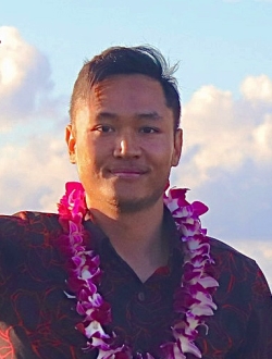 Man with short dark hair wearing a dark red shirt and lei with blue sky and clouds in the background.