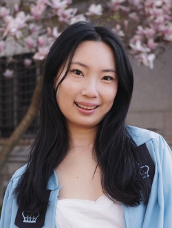 Smiling woman with long black hair wearing a light blue shirt over a white top with a pink flowering tree in the background.