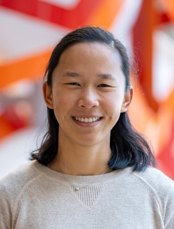Smiling woman with long straight black hair wearing a light colored sweater with a colorful orange and yellow sculpture in the background..