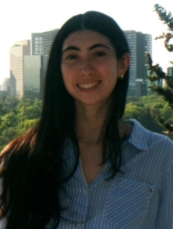 Smiling woman with long dark hair wearing a blue shirt with trees and buildings in the background.
