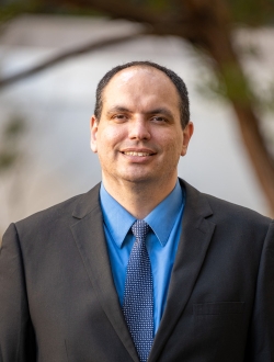 Smiling man with short black hair wearing a dark suit with a blue tie and shirt with a tree and building in the background.