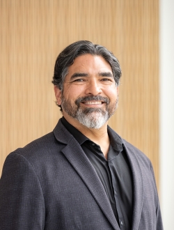 Man with dark hair and a salt and pepper beard wearing a dark gray suit and black shirt.
