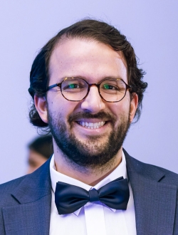Smiling man dark hair and beard wearing glasses and a bow tie.